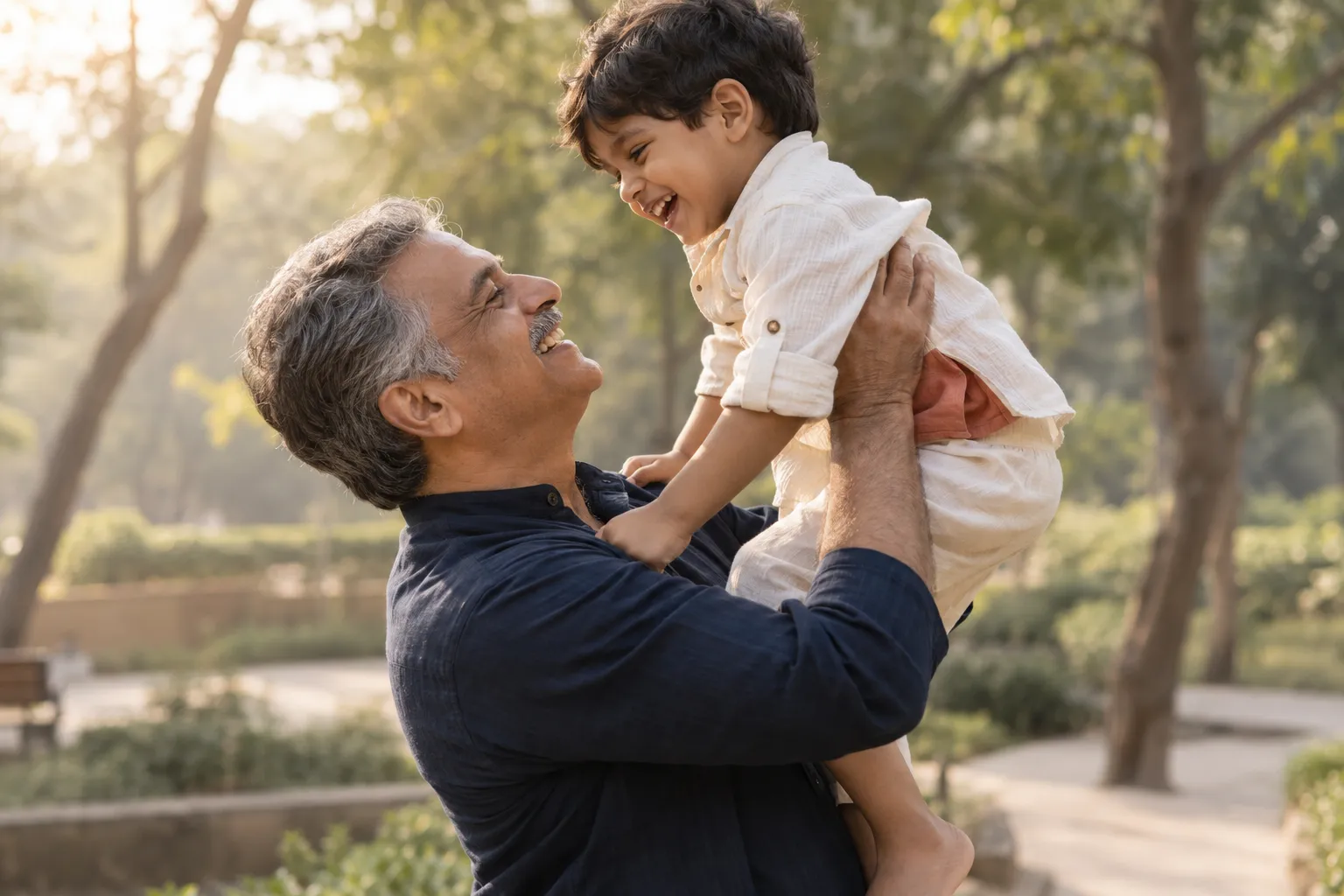 A grandfather in a navy kurta lifts his laughing grandson in a sunlit park, the two smiling at each other.